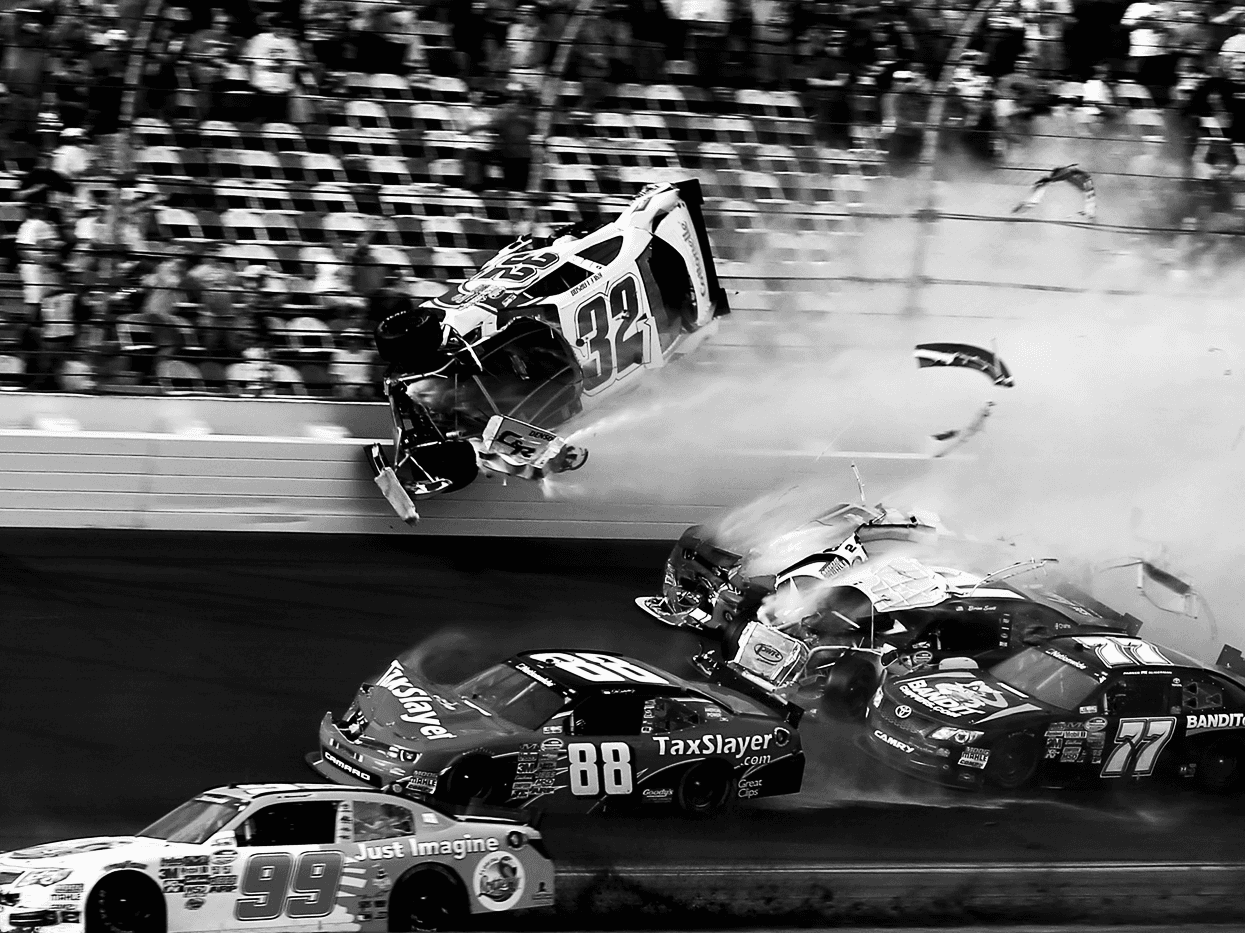 Dramatic black-and-white NASCAR race action photograph with cars, smoke, and grandstand spectators filling the frame